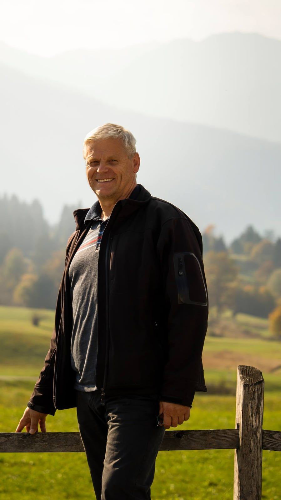 Jože Sodja, nasmejan, ob leseni ograji na travniku nad Bohinjem, v ozadju gozd in megleni vrhovi