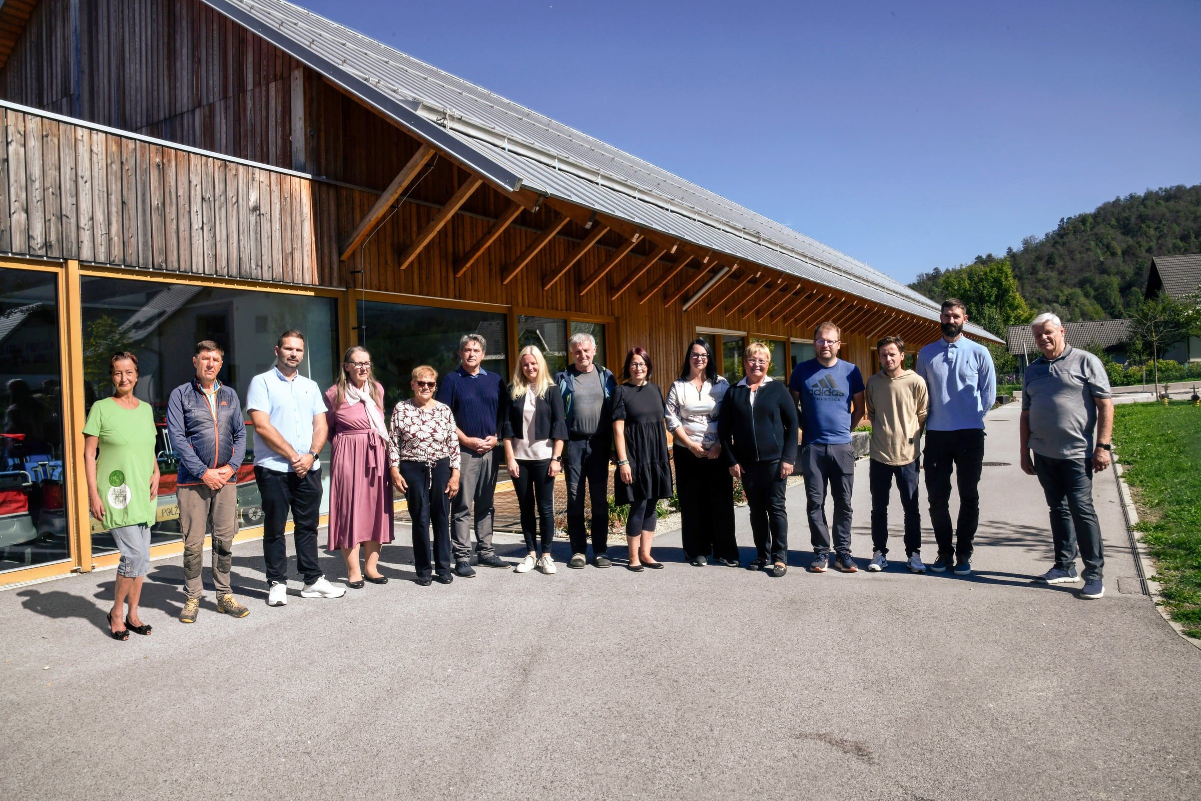 Petnajst kandidatk in kandidatov Liste za Bohinj stoji pred novim vrtcem, leseno stavbo z macesnovo fasado, pod jasnim nebom